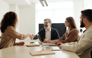A business partnership handshake between two professionals in formal attire, with two colleagues sitting beside them.